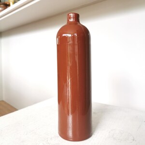 May include: A tall, cylindrical vase in a rich, reddish-brown hue. The ceramic vase has a glossy finish and a narrow neck. The vase is standing on a white surface. The background is a white wall and a white shelf.