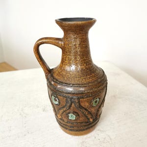 May include: A brown ceramic pitcher with a handle and a textured surface. The pitcher has a dark rim and is decorated with black and green geometric patterns around the base. The pitcher is likely vintage or antique.