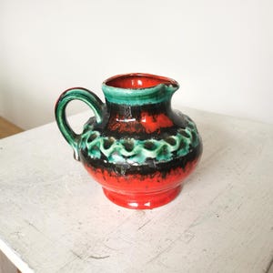 May include: A small, ceramic pitcher with a handle, featuring a vibrant color scheme. The base and rim are red, with a black and green band around the middle. The handle is green, and the pitcher has a glossy finish.