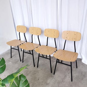 May include: Four wooden chairs with light brown seats and backs, and black metal frames. The chairs are arranged in a row against a white backdrop. The chairs have a simple, minimalist design.
