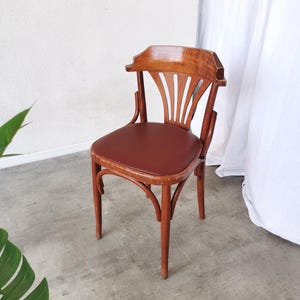 May include: A vintage wooden chair with a curved back and a reddish-brown seat. The chair has a light brown finish and a classic design, suitable for dining or as a decorative piece. The chair's seat is made of a reddish-brown material.