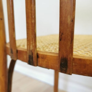 May include: Close-up of the back of a wooden chair with a woven cane seat. The chair has a simple design with vertical slats and a rounded back.