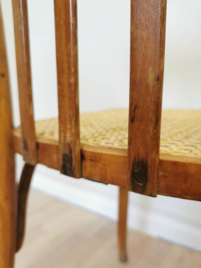May include: Close-up of the back of a wooden chair with a woven cane seat. The chair has a simple design with vertical slats and a rounded back.