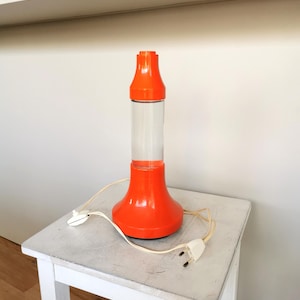 Könnte beinhalten: Orangefarbene Lavalampe mit einem durchsichtigen Glasrohr und einem weißen Kabel. Die Lampe steht auf einem weißen Hocker.