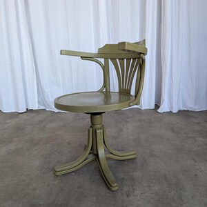 May include: An olive green vintage desk chair with a round seat and curved back. The chair has a small armrest and a pedestal base with curved legs. The chair is made of wood and has a distressed finish.