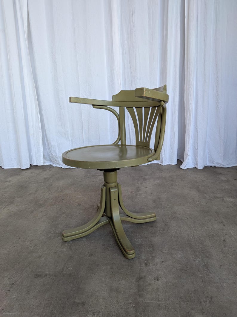 May include: An olive green vintage desk chair with a round seat and curved back. The chair has a small armrest and a pedestal base with curved legs. The chair is made of wood and has a distressed finish.