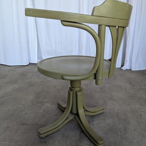 May include: A vintage olive green office chair with a curved back and armrest. The chair has a round seat and a pedestal base with four legs. The chair is made of wood and has a distressed finish.