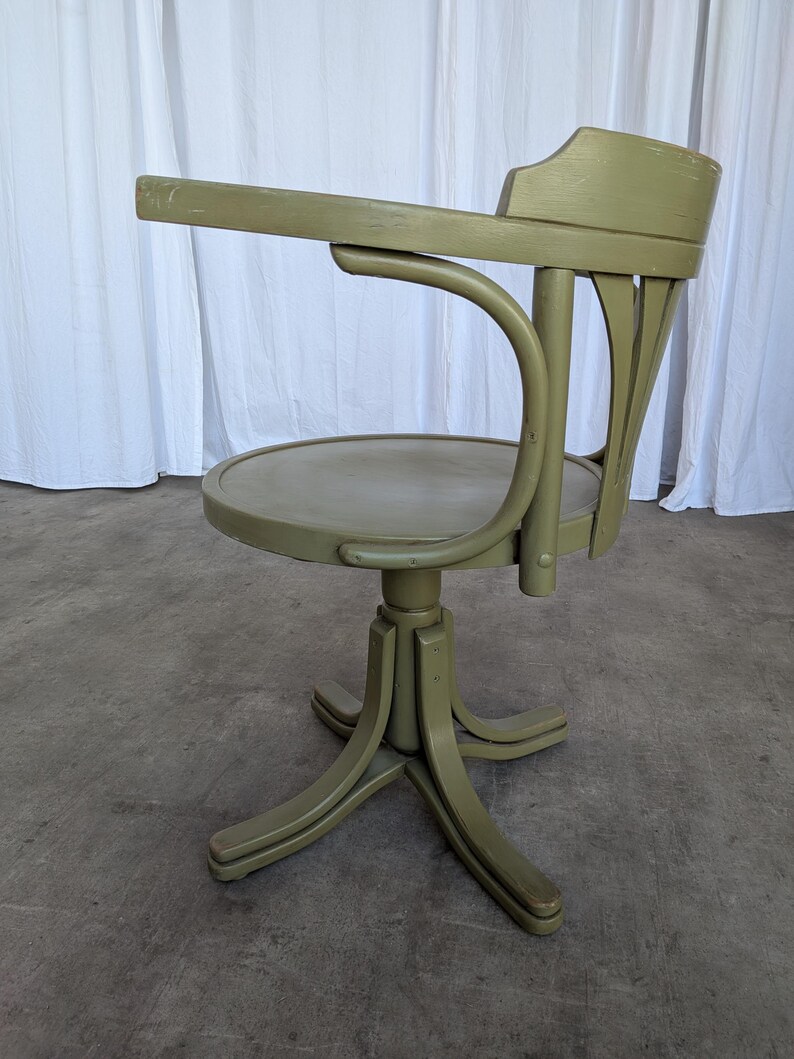 May include: A vintage olive green office chair with a curved back and armrest. The chair has a round seat and a pedestal base with four legs. The chair is made of wood and has a distressed finish.