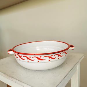 May include: White enamel bowl with red floral design and two handles. The bowl is sitting on a white wooden stool.