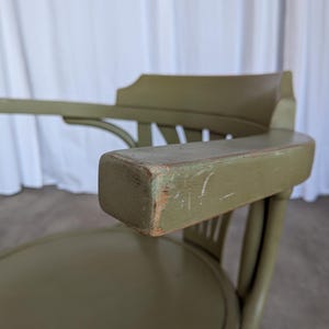May include: Close-up of an olive green wooden chair with armrests. The chair has a rounded seat and a curved back. The armrest shows signs of wear, with visible scratches and exposed wood.