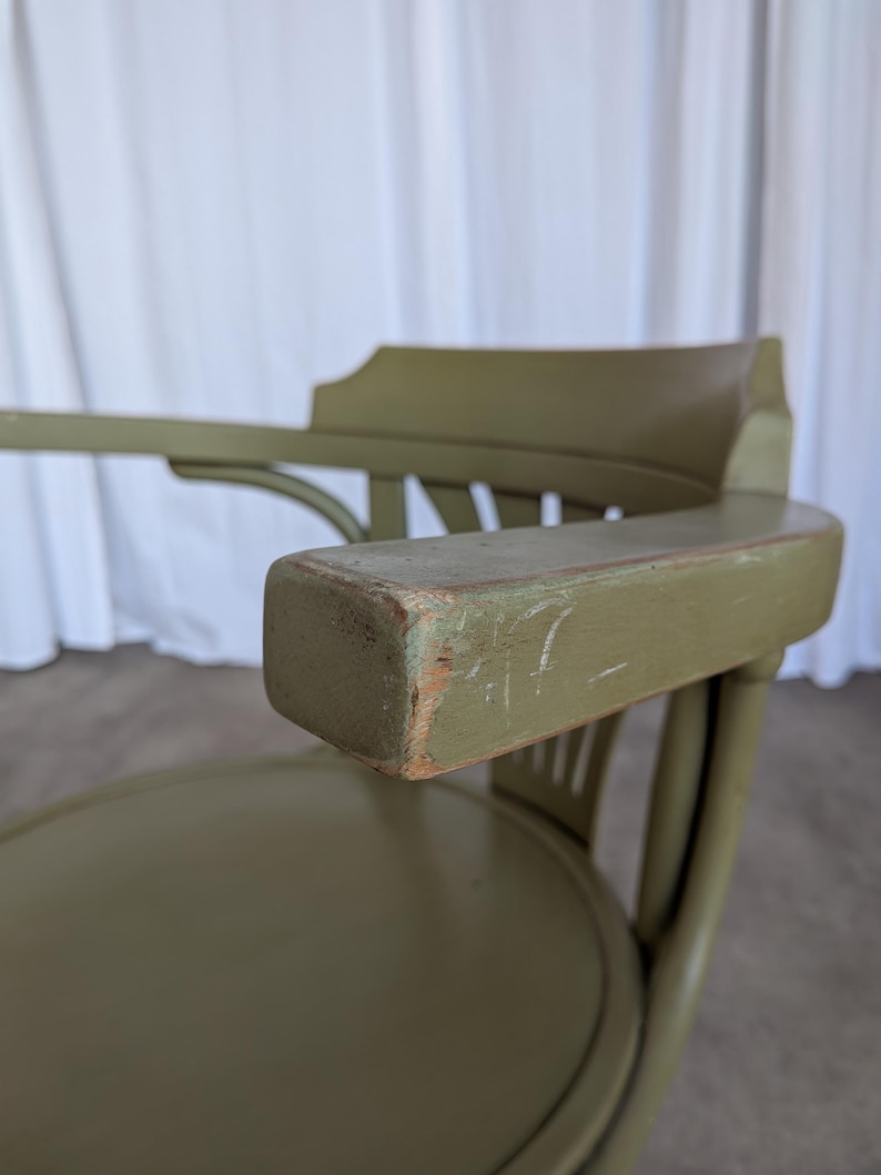 May include: Close-up of an olive green wooden chair with armrests. The chair has a rounded seat and a curved back. The armrest shows signs of wear, with visible scratches and exposed wood.