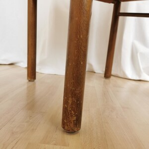 May include: Close-up of the wooden leg of a chair. The leg is brown and has a worn finish. The chair is sitting on a light wood floor.