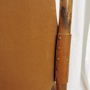 May include: A brown canvas chair with a wooden frame and leather accents. The chair has a unique design with a curved back and a wide seat. The leather accents are secured with brass tacks.