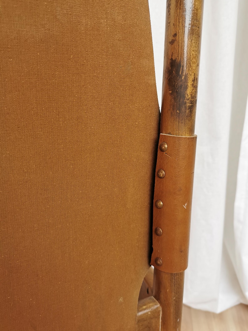 May include: A brown canvas chair with a wooden frame and leather accents. The chair has a unique design with a curved back and a wide seat. The leather accents are secured with brass tacks.