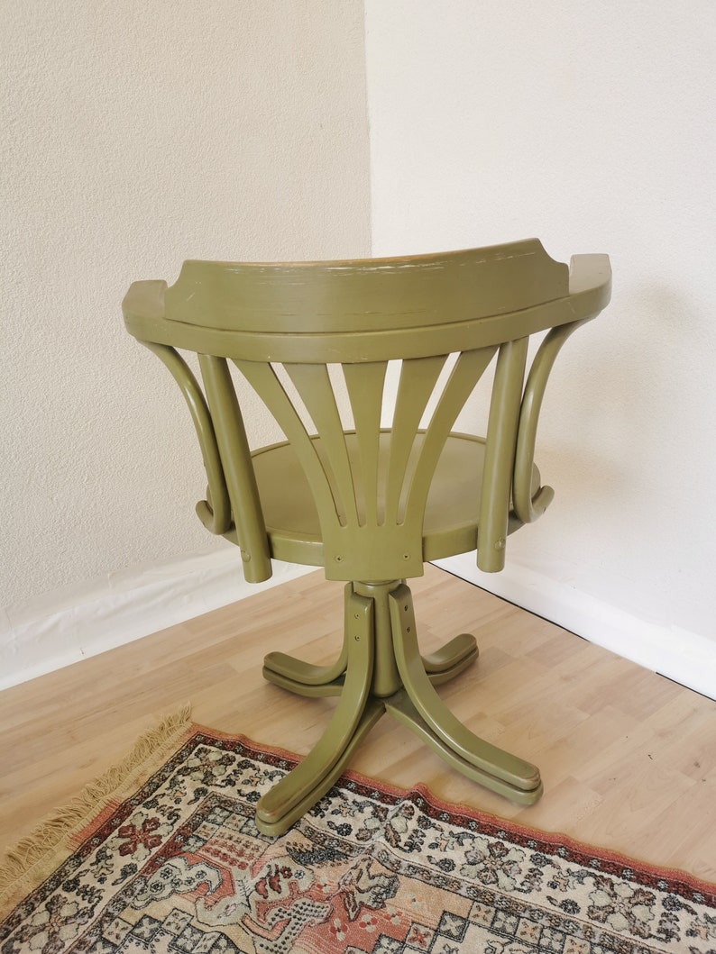 May include: A vintage olive green swivel chair with a curved back and a round seat. The chair has a star-shaped base and is positioned in a corner. The chair is made of wood and has a distressed finish.