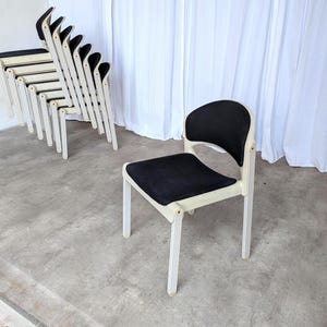 May include: A stack of chairs with off-white frames and black upholstered seats and backrests. Each chair features a curved backrest design and four straight legs. The chairs are arranged with one standing alone in the foreground and several others stacked behind it in a row.