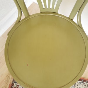 May include: A top-down view of a vintage, olive green chair. The chair has a round seat and a curved back with vertical slats. The chair's frame is a matching olive green. The chair is set on a light wood floor with a patterned rug.