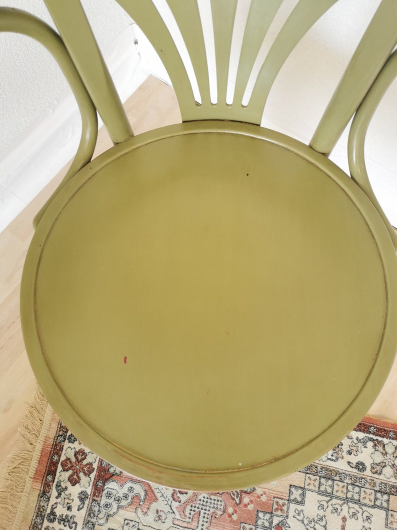 May include: A top-down view of a vintage, olive green chair. The chair has a round seat and a curved back with vertical slats. The chair's frame is a matching olive green. The chair is set on a light wood floor with a patterned rug.