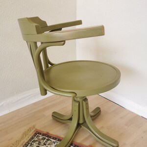 May include: An olive green vintage office chair with a round seat and curved back. The chair has armrests and a five-point base. The chair is made of wood and has a swivel function.
