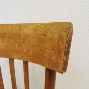 May include: Close-up of the top of a wooden chair with a light brown finish. The chair has a curved back and vertical slats.