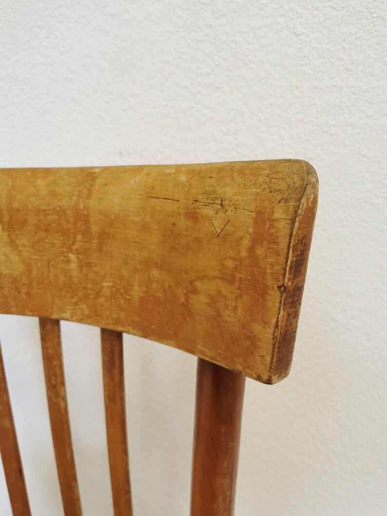 May include: Close-up of the top of a wooden chair with a light brown finish. The chair has a curved back and vertical slats.
