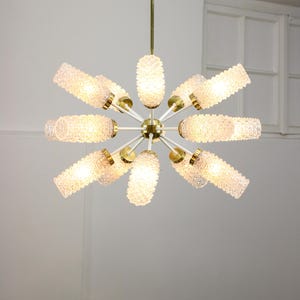 Może przedstawiać: Kryształowy żyrandol z mosiądzu z 12 matowymi szklanymi kloszami. Klosze są cylindryczne i mają fakturowaną powierzchnię. Żyrandol ma biały montaż sufitowy.