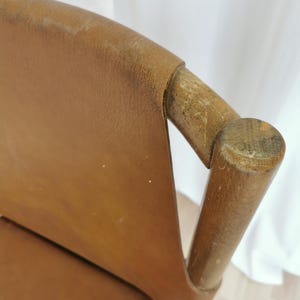 May include: Close-up of a brown leather chair with wooden arms and a wooden back. The chair is in good condition, with some wear and tear on the leather.