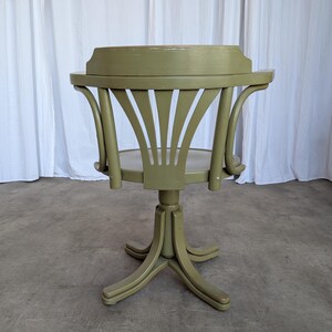 May include: A vintage, olive green wooden chair with a round seat and a curved back. The chair has a pedestal base with four legs and a swivel mechanism. The back of the chair features a fan-like design. The chair is painted in a matte finish.
