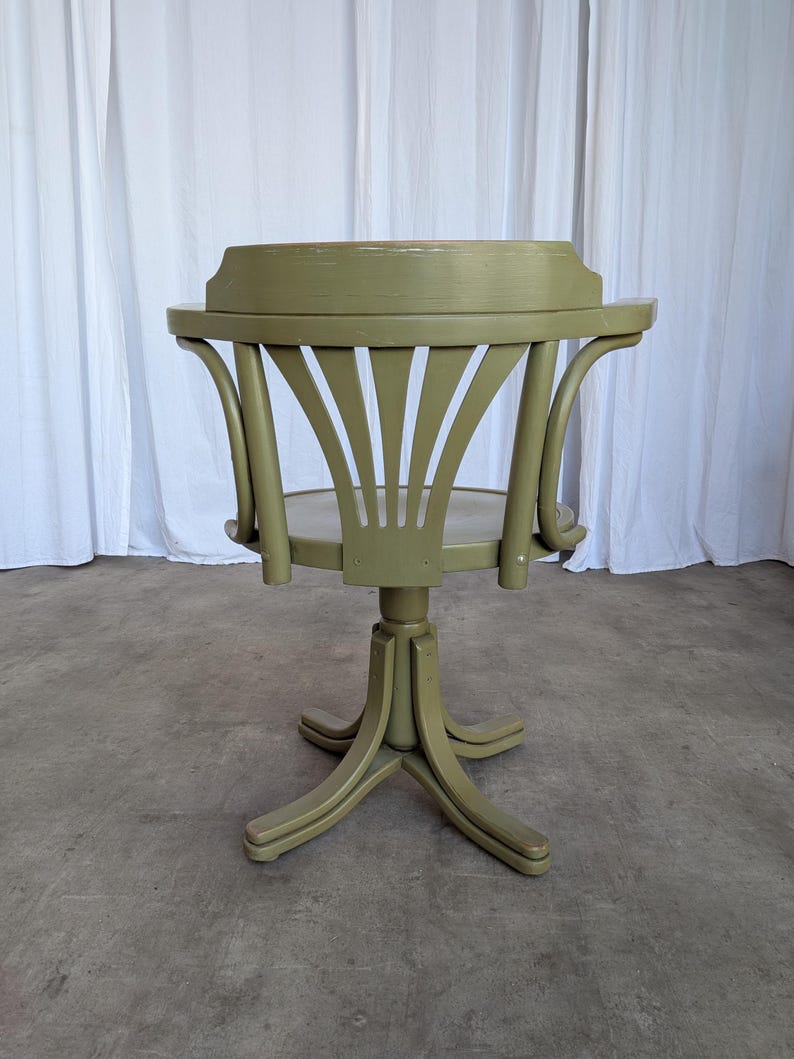 May include: A vintage, olive green wooden chair with a round seat and a curved back. The chair has a pedestal base with four legs and a swivel mechanism. The back of the chair features a fan-like design. The chair is painted in a matte finish.