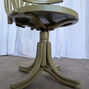 May include: Olive green vintage office chair with a rounded seat and a slatted back. The chair has curved armrests and a pedestal base with four curved legs. The chair is made of wood and has a distressed finish.