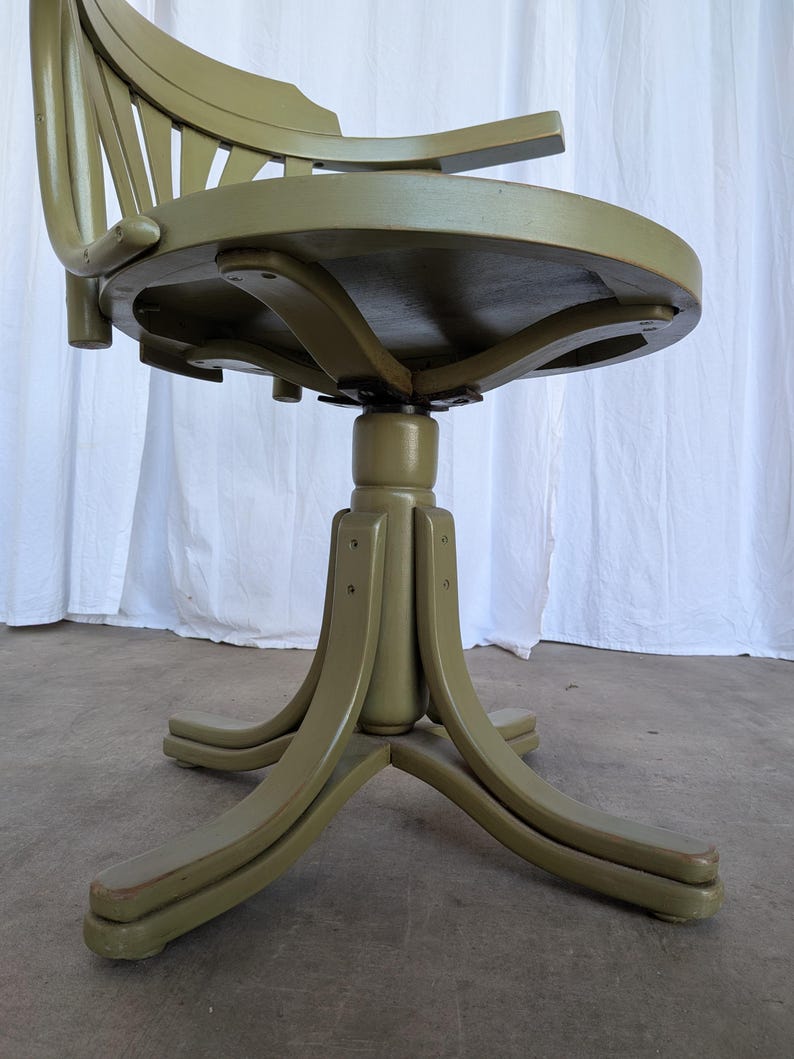 May include: Olive green vintage office chair with a rounded seat and a slatted back. The chair has curved armrests and a pedestal base with four curved legs. The chair is made of wood and has a distressed finish.