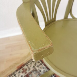 May include: A sage green wooden chair with a curved armrest. The chair has a round seat and a back with a fan-like design. The armrest shows signs of wear, with visible scratches and exposed wood.