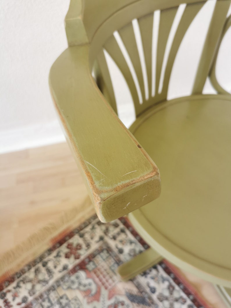May include: A sage green wooden chair with a curved armrest. The chair has a round seat and a back with a fan-like design. The armrest shows signs of wear, with visible scratches and exposed wood.