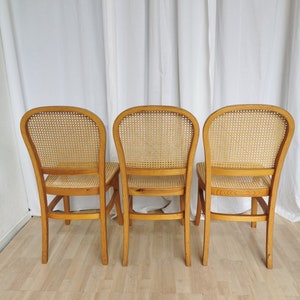 May include: Three wooden chairs with cane seats and backs. The chairs are light brown and have a simple design. They are arranged in a row, facing away from the camera.