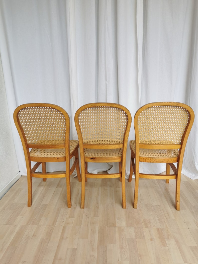 May include: Three wooden chairs with cane seats and backs. The chairs are light brown and have a simple design. They are arranged in a row, facing away from the camera.
