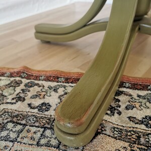 May include: Close-up of a green wooden chair leg. The chair leg has a curved design and is painted in a muted green color. The chair leg is resting on a patterned rug and a wooden floor.