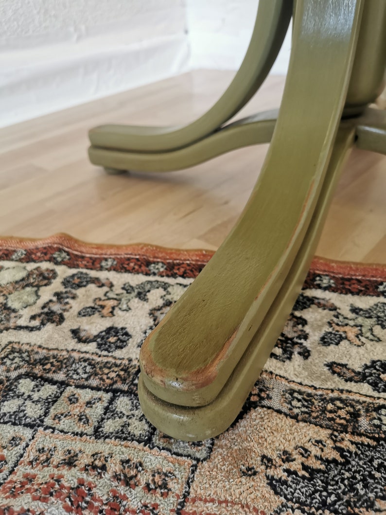 May include: Close-up of a green wooden chair leg. The chair leg has a curved design and is painted in a muted green color. The chair leg is resting on a patterned rug and a wooden floor.