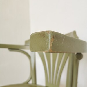 May include: Close-up of a green wooden chair with a curved back and armrest. The chair has a distressed finish, revealing the wood underneath in some areas. The design features vertical slats on the back and a rounded seat.