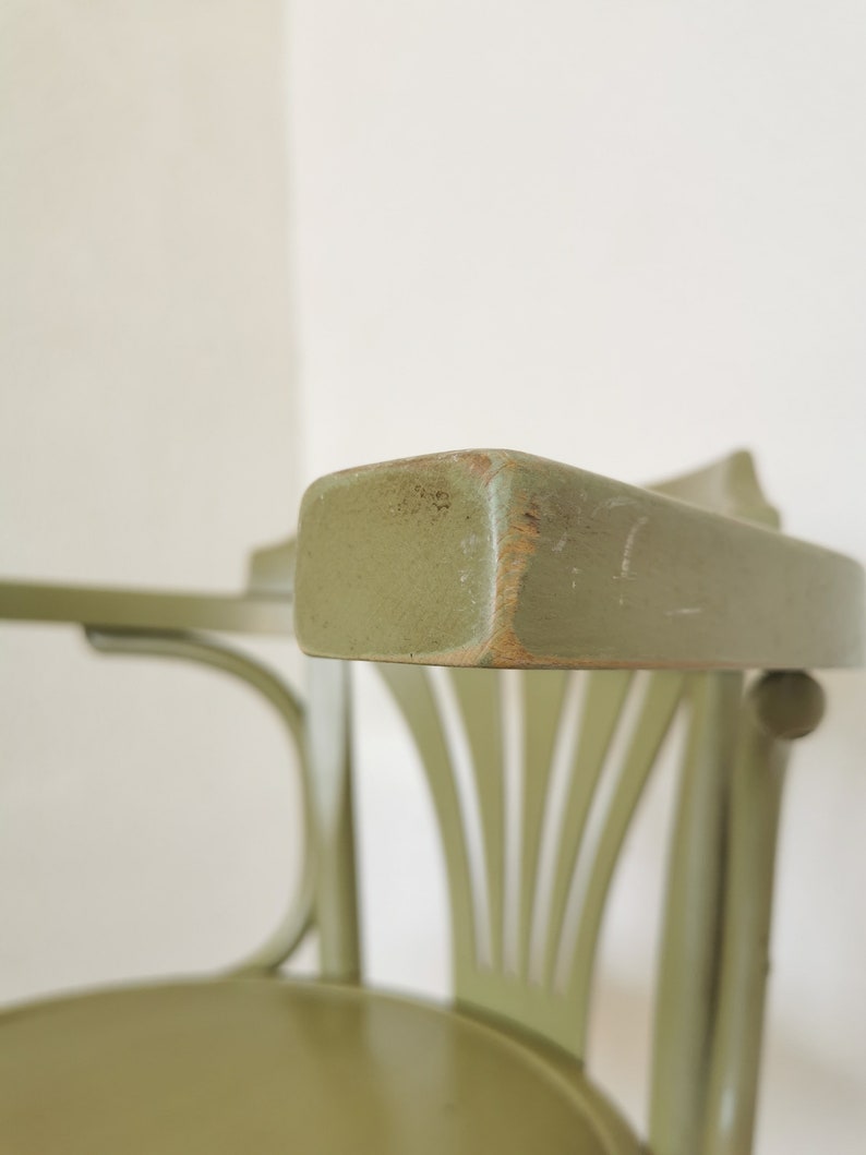 May include: Close-up of a green wooden chair with a curved back and armrest. The chair has a distressed finish, revealing the wood underneath in some areas. The design features vertical slats on the back and a rounded seat.