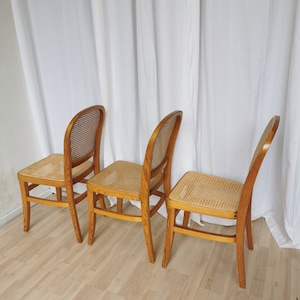 May include: Three wooden chairs with cane seats. The chairs are light brown and have a simple design. The chairs are arranged in a row on a wooden floor.