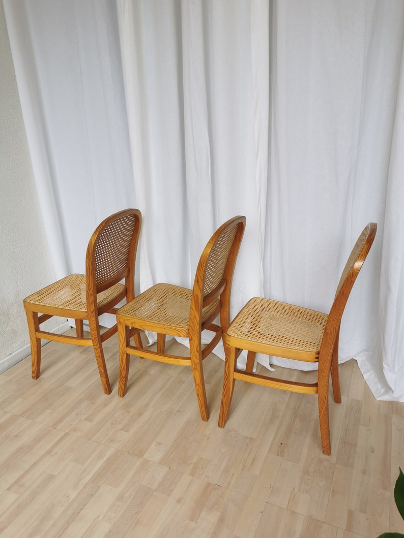 May include: Three wooden chairs with cane seats. The chairs are light brown and have a simple design. The chairs are arranged in a row on a wooden floor.