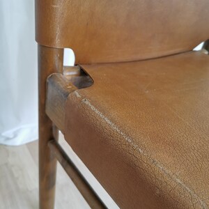 May include: Close-up of a brown leather chair with a wooden frame. The chair has a simple design with a slightly curved back and a wide seat. The leather is worn and has a distressed look.