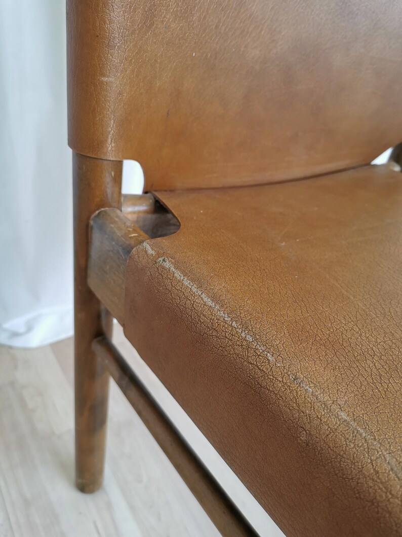 May include: Close-up of a brown leather chair with a wooden frame. The chair has a simple design with a slightly curved back and a wide seat. The leather is worn and has a distressed look.
