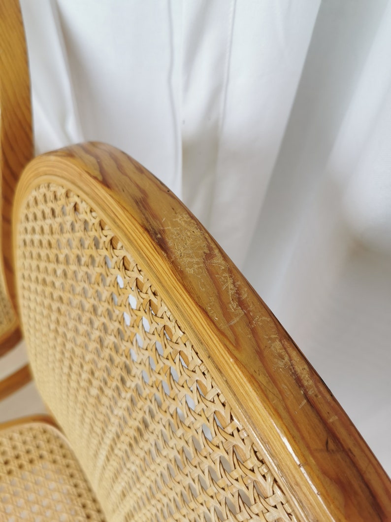May include: Close-up of a wooden chair with a woven cane seat and back. The chair is made of light brown wood and has a natural finish. The cane is woven in a tight pattern and is in good condition.