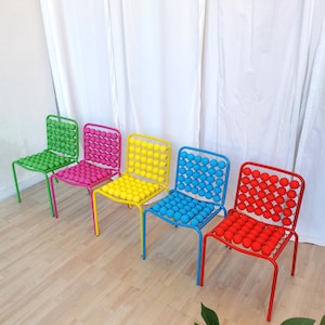 May include: Five colorful chairs with a unique design. The chairs are made of metal and have a seat made of small, round balls. The chairs are arranged in a row, with the green chair on the left and the red chair on the right. The chairs are all different colors: green, pink, yellow, blue, and red.