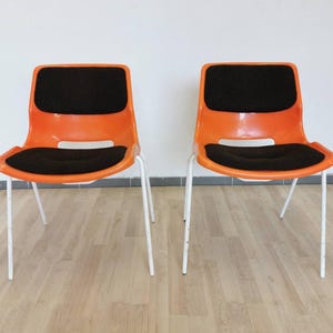 May include: Two orange and black chairs with white metal legs. The chairs have a simple, modern design.