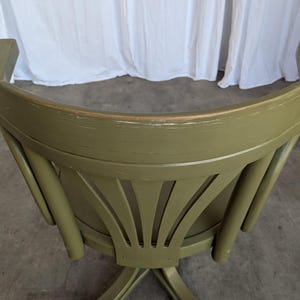 May include: An olive green wooden chair with a curved back and decorative fan-shaped cutouts. The chair has a distressed finish, and the legs are crossed. The background is a white curtain.