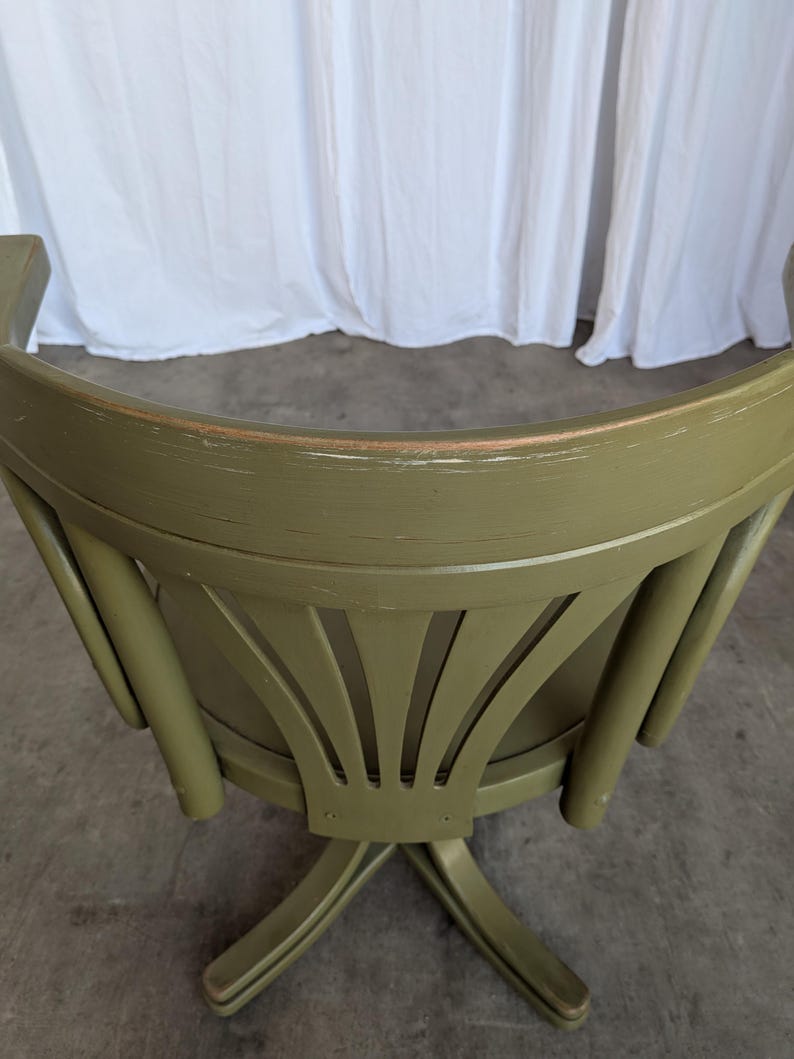 May include: An olive green wooden chair with a curved back and decorative fan-shaped cutouts. The chair has a distressed finish, and the legs are crossed. The background is a white curtain.