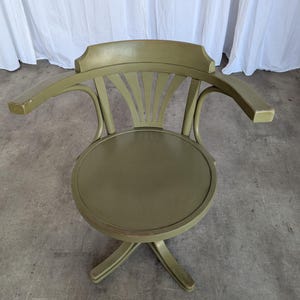 May include: An olive green wooden swivel chair with armrests. The chair has a round seat and a curved back with a fan-like design. The chair's legs are in a star-like formation. The chair is painted in a muted green color.