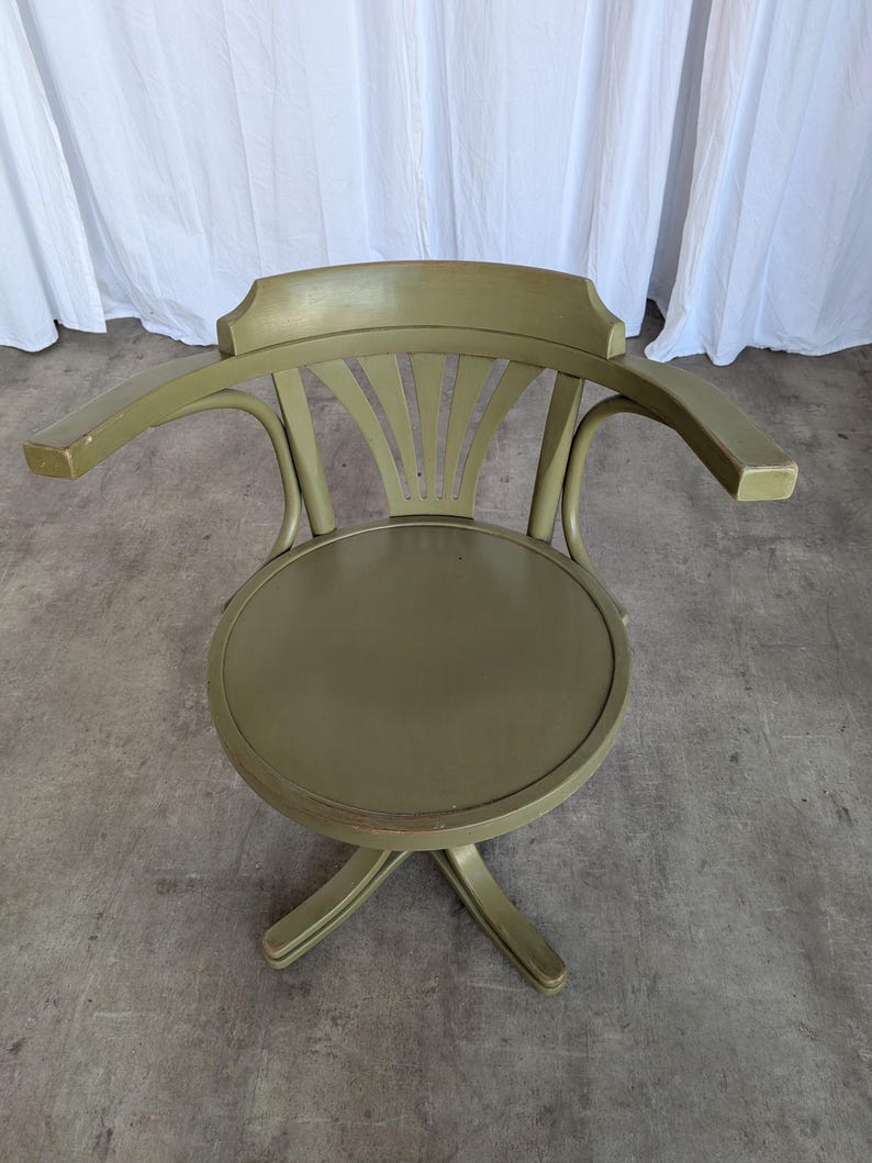 May include: An olive green wooden swivel chair with armrests. The chair has a round seat and a curved back with a fan-like design. The chair's legs are in a star-like formation. The chair is painted in a muted green color.